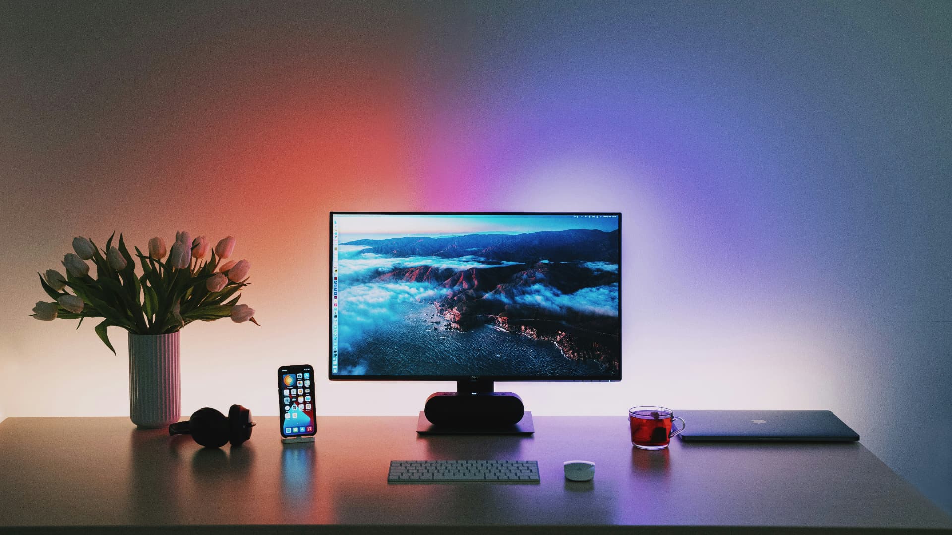 Een verzorgd bureau met een computer, toetsenbord, koptelefoon en een vaas tulpen